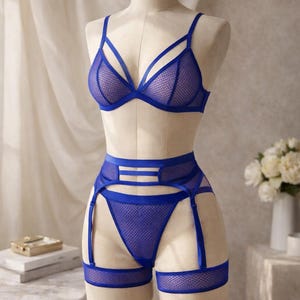 May include: A blue lingerie set displayed on a mannequin. The set includes a triangle bra, high-waisted briefs, and a garter belt. The lingerie is made of a mesh material with blue straps and trim. The mannequin is on a gold stand.