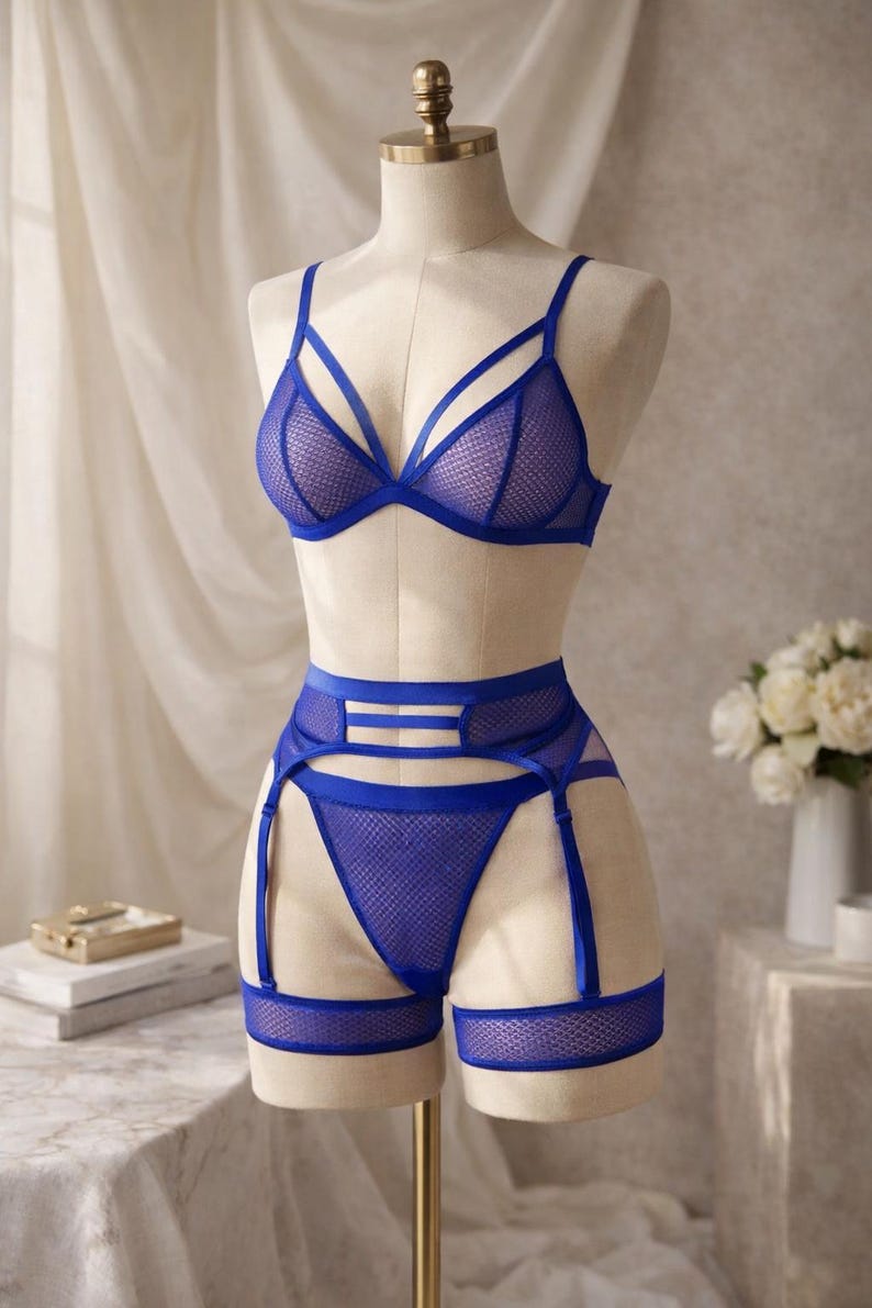 May include: A blue lingerie set displayed on a mannequin. The set includes a triangle bra, high-waisted briefs, and a garter belt. The lingerie is made of a mesh material with blue straps and trim. The mannequin is on a gold stand.