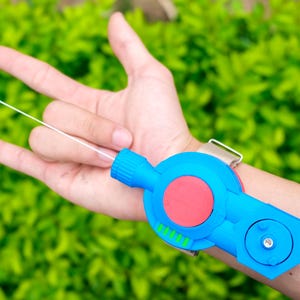 May include: A blue wrist-mounted toy shooter featuring a prominent red circular button in the center and a small nozzle emitting a thin white string. The device is secured to the wrist with a metallic strap and includes a secondary circular component.