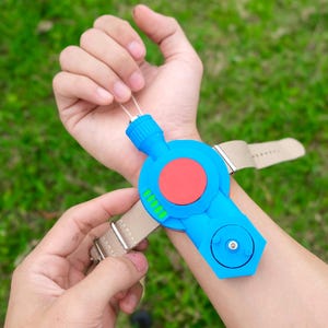May include: A blue wrist-mounted device featuring a large red circular button, a smaller circular dial, and four green indicator lights. The device is attached to a beige fabric strap with a silver metal buckle, secured around a wrist.