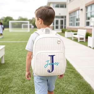May include: Cream-colored quilted backpack with a front pocket and a top handle. The backpack features a large navy blue letter "J" and the name "James" embroidered in teal. The backpack is worn by a child in a light blue shirt and shorts.