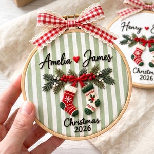 May include: Two round wooden embroidery hoops featuring fabric backgrounds, one with green and white stripes and one with solid white. Both display embroidered Christmas stockings, pine branches, red bows, and the names Amelia, James, Henry, and Evelyn.