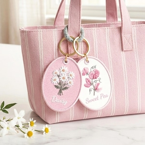 Peut inclure: Un sac fourre-tout rose à rayures blanches avec deux porte-clés floraux. Un porte-clés présente un motif de marguerite, et l'autre un motif de pois de senteur. Les porte-clés sont attachés à la poignée du sac avec des anneaux dorés.