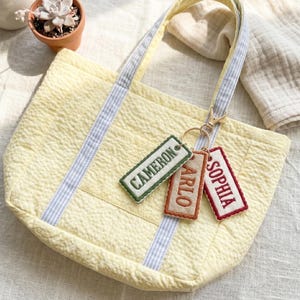 May include: A yellow quilted tote bag with blue and white striped handles. Three rectangular keyrings are attached, embroidered with the names "CAMERON", "ARLO", and "SOPHIA". The bag rests on a white surface.