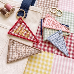 May include: A collection of triangular fabric keychains featuring gingham patterns in red, blue, pink, green, and brown. Each keychain is embroidered with names including Jack, Harry, Sophia, and Maya, and includes a gold-toned metal keyring.