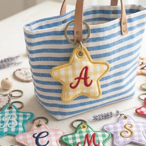 Peut inclure: Un sac fourre-tout à rayures bleues et blanches avec des poignées en cuir et un grand anneau métallique tenant un porte-clés en forme d'étoile à carreaux jaunes. Le porte-clés porte la lettre "A" brodée en rouge. D'autres porte-clés en forme d'étoile avec différentes lettres sont visibles.