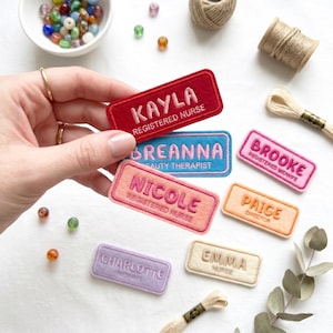 May include: Embroidered name badges in various colours, including red, blue, and pink, with names and job titles such as "Kayla, Registered Nurse" and "Brooke, Registered Midwife." The tags are rectangular with rounded corners.