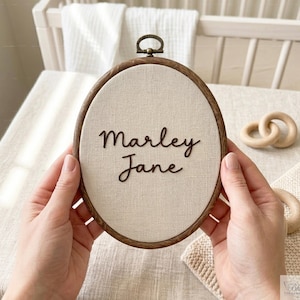 May include: Oval embroidery hoop with the name "Marley Jane" stitched in brown thread on a cream fabric. The hoop has a dark wood finish and a metal hanging loop. The background includes a cot and wooden toys.