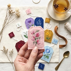 May include: Embroidered felt tea bag tags in various colours, including pink, purple, and blue, each with a white string. The tags feature floral designs. A cup of tea with star anise is in the background.