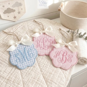 May include: Three embroidered baby name tags with scalloped edges and white bows. One is light blue gingham with blue initials, and two are pink, one with gingham and the other with stripes, each with pink initials. A banner with clouds and stars is in the background.