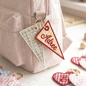 May include: Pink striped backpack with two personalized pennant keychains. One keychain is a gray and white checkered pennant with the name "Maya" embroidered in white script. The other is a yellow and red checkered pennant with the name "Adree" embroidered in red script.