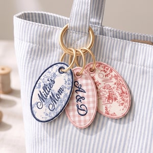 May include: Three oval-shaped fabric bag tags attached to a gold-toned metal ring on a light blue and white striped tote bag. The tags feature embroidered text including Millie's Mom, A & D, and Maman, with blue floral, pink gingham, and red floral patterns.