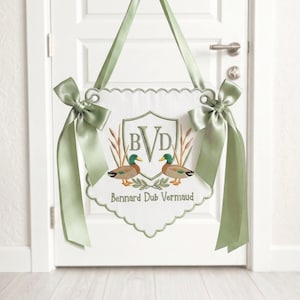 May include: A white door hanging with a scalloped edge, featuring embroidered ducks and the initials "BVD" within a crest. The design includes the text "Bennard Dub Vermaud" and is accented with sage green bows and ribbons.