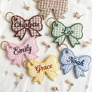 May include: A collection of six fabric bow keychains featuring a gingham pattern in brown, green, pink, yellow, and blue. Each bow has a gold-toned metal keyring and is embroidered with a name: Olivia, Charlotte, Leo, Emily, Grace, and Noah.