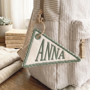 May include: A personalized keychain with the name "ANNA" embroidered in green on a white pennant. The pennant has a green scalloped border and is attached to a gold keyring. The keychain hangs from a striped backpack.
