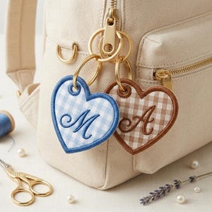 May include: Two heart-shaped fabric keychains attached to a beige canvas backpack. One heart features a blue gingham pattern with an embroidered letter M, and the other features a brown gingham pattern with an embroidered letter A in dark brown.