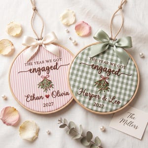 May include: Two circular wooden embroidery hoops featuring fabric with embroidered text THE YEAR WE GOT engaged, a mistletoe sprig, names, and the year 2027. One hoop has pink stripes and a white bow, the other green gingham and a sage green bow.