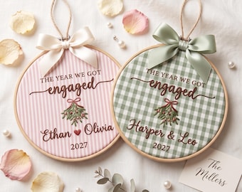 Peut inclure: Deux tambours à broder circulaires en bois avec du tissu brodé portant le texte THE YEAR WE GOT engaged, un brin de gui, des prénoms et l'année 2027. Un tambour a des rayures roses et un nœud blanc, l'autre un vichy vert et un nœud vert sauge.