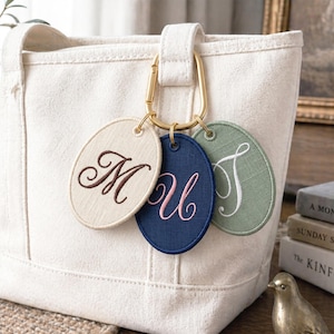 May include: A cream-colored tote bag with a gold carabiner clip holding three embroidered oval tags. The tags display the letters "M", "U", and "J" in brown, pink, and white thread, respectively. The bag is made of a textured fabric.