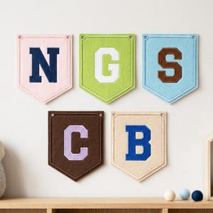 May include: Five felt pennant banners in pastel shades, each displaying a different letter: N, G, S, C, and B. The letters are in a contrasting colour and stitched onto the pennants. The banners are arranged on a white wall above a wooden shelf.