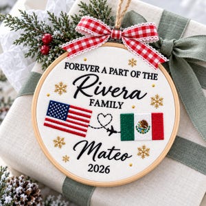 May include: A circular embroidery hoop ornament featuring white fabric with the embroidered text FOREVER A PART OF THE Rivera FAMILY, Mateo, and 2026. It includes a US flag, a Mexican flag, a dashed flight path with a heart and plane, and gold snowflakes.