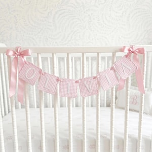 May include: A decorative banner featuring seven scalloped, light pink and white gingham fabric squares, each embroidered with a white letter spelling OLIVIA. The banner is finished with a light pink satin ribbon bow on each end.