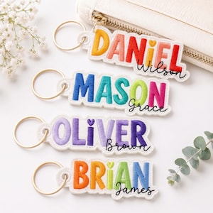May include: Four embroidered fabric keychains with gold-tone metal rings. Each features a name in large, colorful, block-style letters with a smaller cursive name underneath. The names are Daniel Wilson, Mason Grace, Oliver Brown, and Brian James.