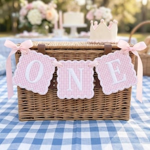 May include: A wicker picnic basket with a banner that spells "ONE" in white letters on pink gingham squares. The banner is decorated with pink ribbons. A small crown sits on top of the basket, and the scene is set on a blue and white checkered tablecloth.