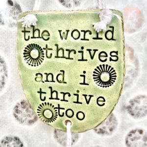 May include: A green ceramic hanging ornament with the text "the world thrives and i thrive too". The ornament has a white tassel and two black and white circular designs.