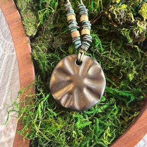 May include: A brown ceramic pendant with a textured surface hangs from a necklace with turquoise and gold beads. The necklace is displayed on a bed of green moss.