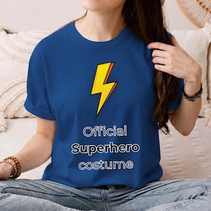 Pode incluir: Uma t-shirt de manga curta azul royal com um gráfico de um raio amarelo com textura de pontos pretos. Abaixo do raio, o texto "Official Superhero costume" está impresso em branco com um contorno preto.