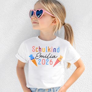 Puede incluir: Una camiseta blanca de manga corta con el texto colorido Schulkind, el nombre Emilia en letra cursiva negra y el año 2026 en azul y rosa. En la parte frontal aparecen dos ilustraciones de conos de regalo con lunares.