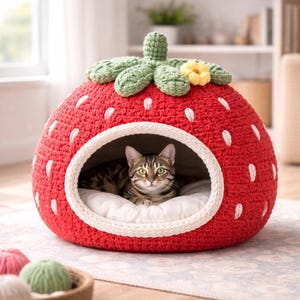 May include: A red crocheted cat bed shaped like a strawberry, with white seed details and green leaves. A cat is inside the bed, which has a white interior cushion and a cream-colored trim around the opening. A yellow flower is on top.