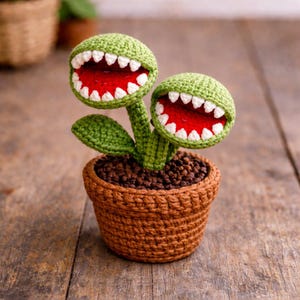 May include: A crocheted plant with two green, round heads, each with a wide, open mouth featuring white teeth and a red interior. The plant sits in a brown crocheted pot filled with brown material, resembling soil. The plant has green stems and leaves.