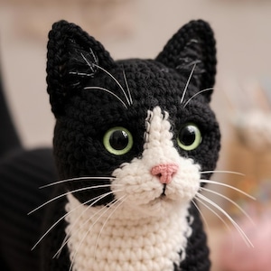 Puede incluir: Primer plano de un gato tejido a ganchillo en blanco y negro, con ojos verdes y nariz rosa. El gato tiene largos bigotes blancos. Las orejas del gato son puntiagudas y el pelaje es texturizado.