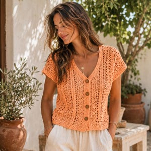 May include: A peach-colored, short-sleeved crochet top with a V-neck and wooden buttons. The open weave design allows for breathability. The top is paired with white pants, creating a casual, summery look. The top is a versatile addition to any wardrobe.