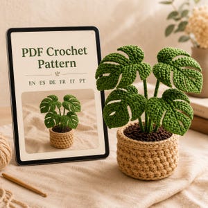 Puede incluir: Una tableta que muestra un patrón de ganchillo en PDF con texto en inglés, español, alemán, francés, italiano y portugués, junto a una planta monstera de ganchillo en una maceta beige con tierra marrón oscura y un ganchillo de madera.
