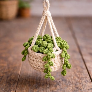May include: A handmade, hanging crochet planter with a cream-colored basket and rope. The basket is filled with green, crocheted succulent plants. The planter is suspended from a knotted rope, creating a charming, rustic aesthetic.