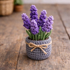 May include: A handmade crochet lavender plant in a gray pot. The lavender flowers are a vibrant purple, with green leaves. The pot is adorned with a twine bow. The craft is displayed on a wooden surface.