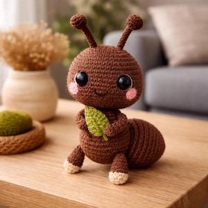 May include: A handcrafted brown crochet ant toy with big black eyes, pink cheeks, and a smiling expression. The ant holds a green leaf and has cream-coloured feet. The toy is placed on a wooden surface.