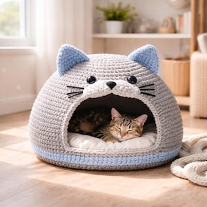 May include: A gray crocheted cat bed shaped like a cat's head, with blue ears and a blue stripe around the base. The bed has a white interior and a sleeping tabby cat inside. Black whiskers and eyes complete the cat face design.