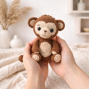 May include: A crocheted monkey doll in shades of brown and cream. The handmade stuffed animal has a round head with large ears, a cream-colored face, and a cream-colored belly. The monkey is being held in two hands.