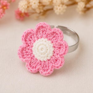 May include: A pink and white crocheted flower ring. The flower has pink petals and a white center, attached to a silver-colored metal ring. The ring is a floral accessory.