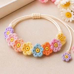 Può includere: Un braccialetto di fiori all'uncinetto colorato con cordino avorio regolabile. Il braccialetto presenta una serie di fiori all'uncinetto nei toni del viola, rosa, arancione, giallo e blu, ciascuno con un centro giallo. Due piccoli ciondoli a forma di fiore pendono alle estremità.
