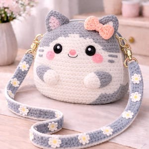 Puede incluir: Bolso de mano con forma de gato tejido a ganchillo, gris y blanco, con un lazo rosa y una correa adornada con margaritas. El bolso tiene cierre de cremallera y enganches de cadena dorados. El gato tiene mejillas rosas y una cara sonriente.