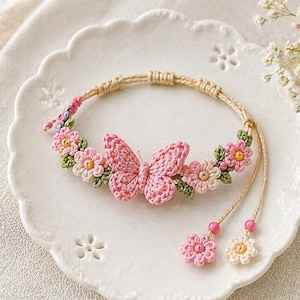 Può includere: Un braccialetto all'uncinetto fatto a mano con un motivo a farfalla e fiori rosa. Il braccialetto è adornato con fiori rosa e crema e foglie verdi. Il braccialetto è esposto su un piatto bianco.