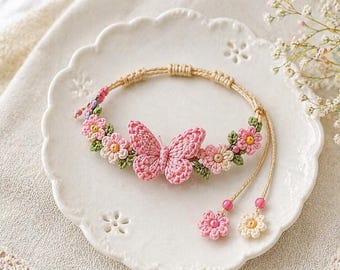 Pode incluir: Uma pulseira de crochê feita à mão com um design de borboleta e flores rosa. A pulseira é adornada com flores rosa e creme e folhas verdes. A pulseira é exibida em um prato branco.