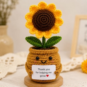 May include: A crocheted sunflower with bright yellow petals and a dark brown centre, sitting in a tan crocheted pot with a smiling face. The pot holds a white rectangular sign that reads, Thank you for helping me grow, with a small red heart.