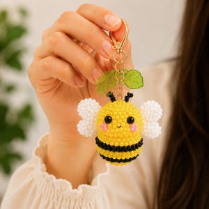 May include: A beaded bee keychain featuring a round yellow body with black horizontal stripes, white beaded wings, and two green translucent leaves attached to a gold-toned metal lobster clasp and ring. The bee has black eyes and pink cheeks.