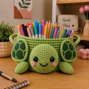 May include: A green crocheted turtle-shaped pen holder filled with colorful pens and pencils. The turtle has a smiling face, black eyes, and pink cheeks. The holder has handles and turtle flipper details.